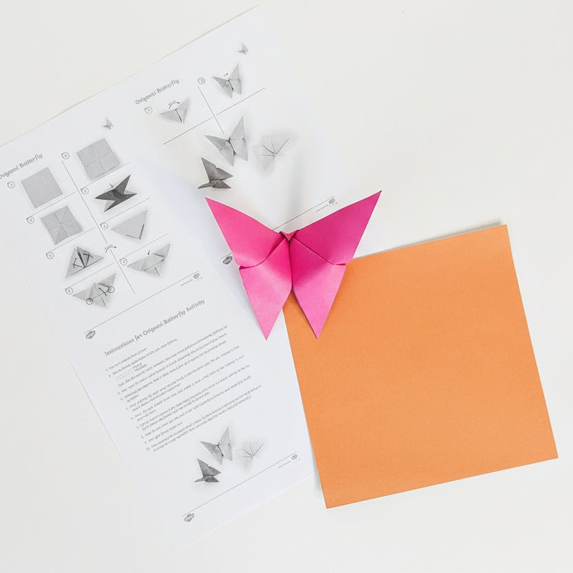 Pink origami butterfly with instructions and orange square paper on a white background