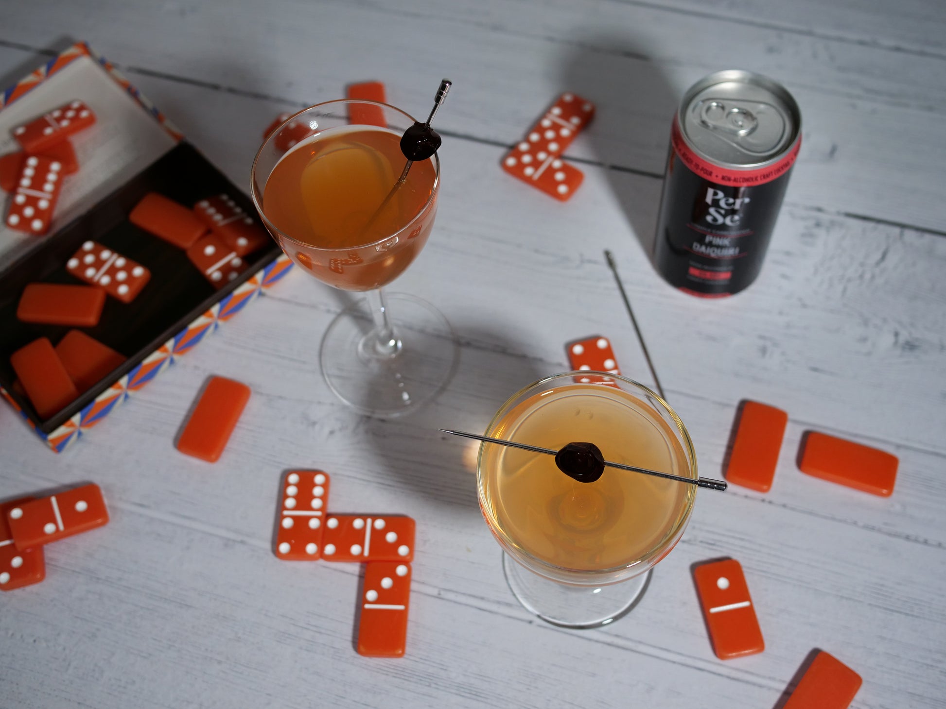 Pink Daiquiri can with two cocktails garnished with cherries with dominoes strewn around.